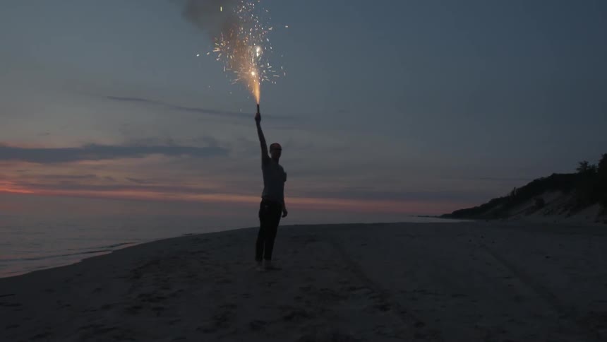 A man holds a burning flare towards the sky as he stands on a beach at dusk