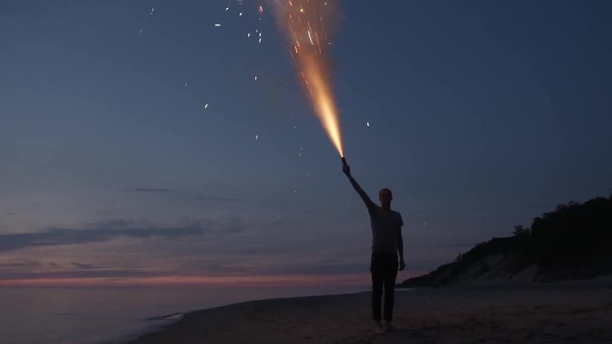 A man standing on a beach holds a flare towards the sky as it burns