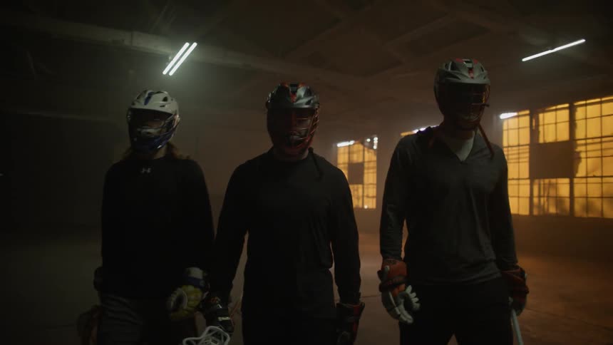 Three people in lacrosse gear walk in a dimly lit warehouse