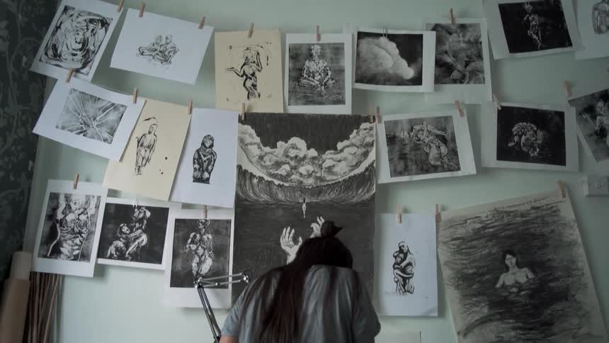 An artist sits at a desk sketching below hanging pictures