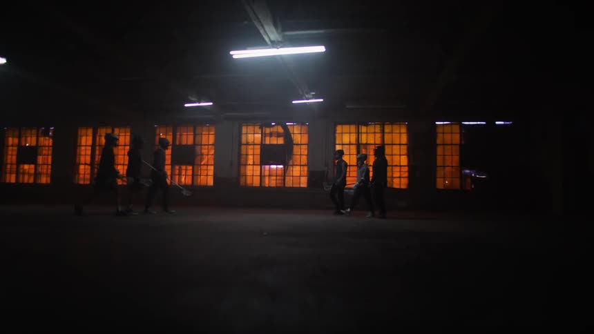 Two teams of people holding lacrosse gear walk up to each other in a dark warehouse