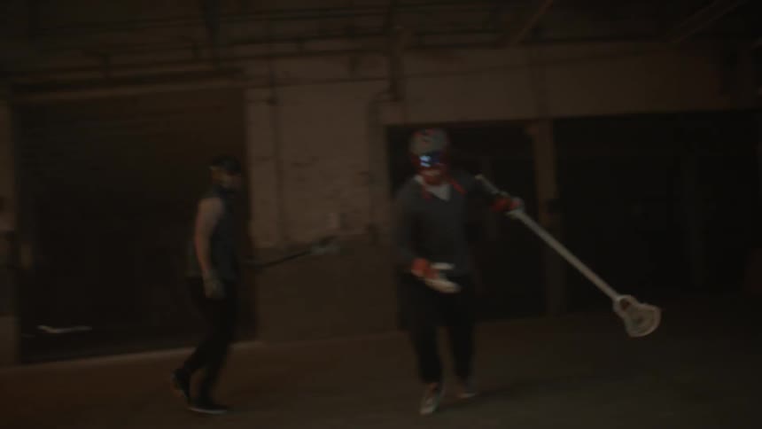 Two people play lacrosse in a dim warehouse