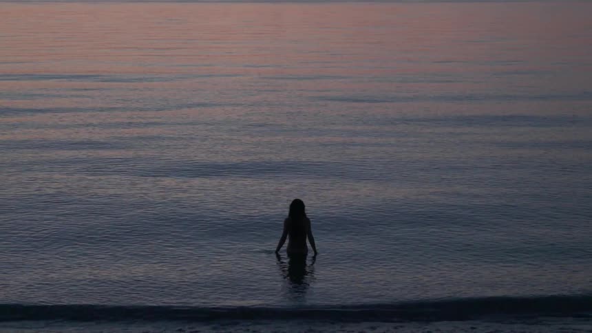 A woman is walking into the ocean at dusk