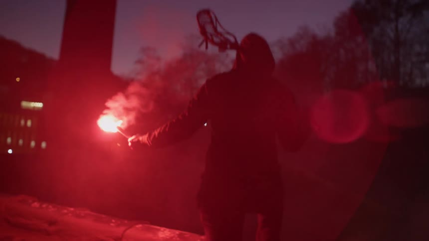 A man holds a lacrosse stick and a red flare