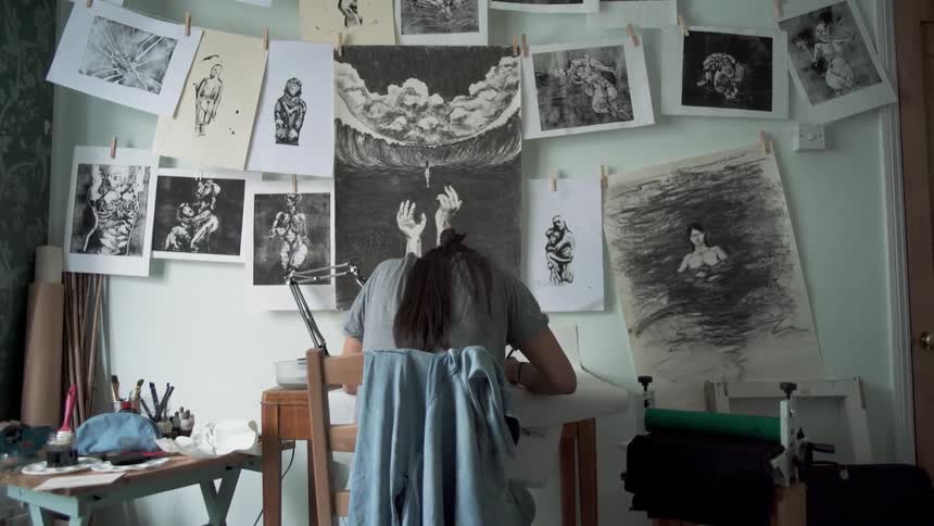 Woman sits at a desk drawing with pictures of drawings hanging up by her