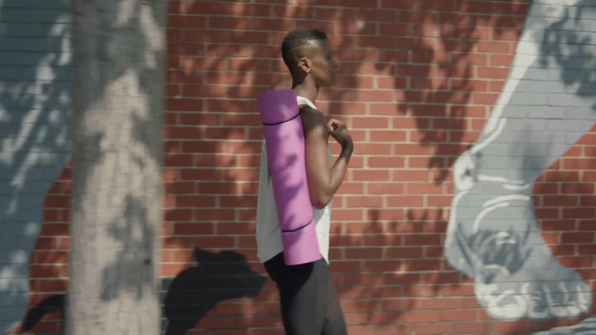 A black woman carrying a yoga mat is walking past a brick wall