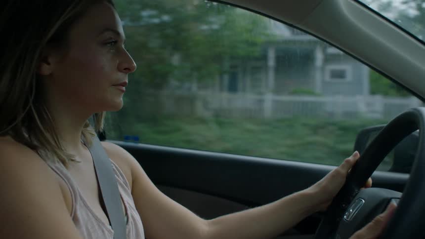 A woman is driving along a row of houses with lots of green trees