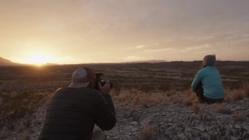 A man photographs a woman in a rural field at sunset