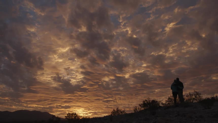 A couple stand on a hill watching the sunset