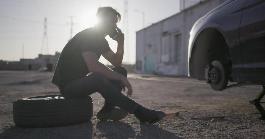 A man sits on a tire and talks on his phone