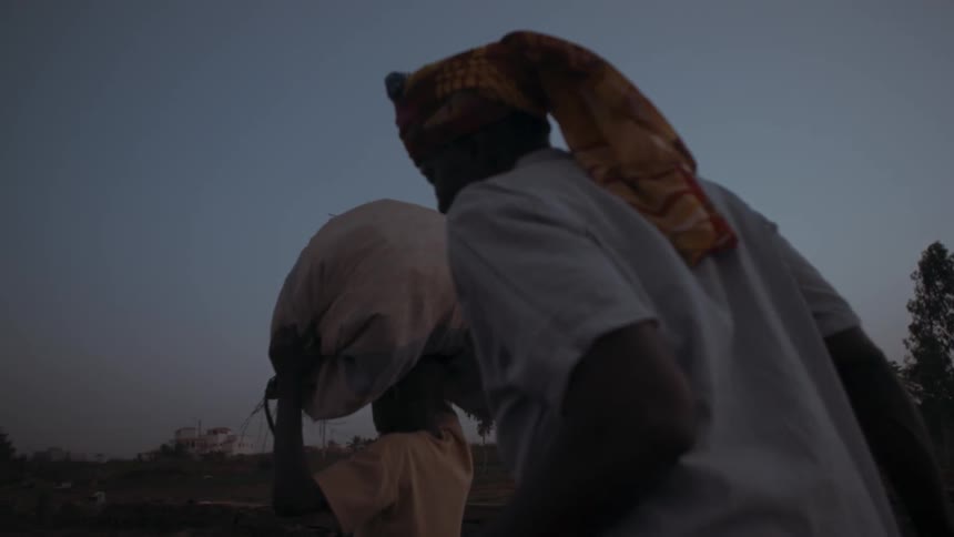 People walking in a field and one carries a large package on their head