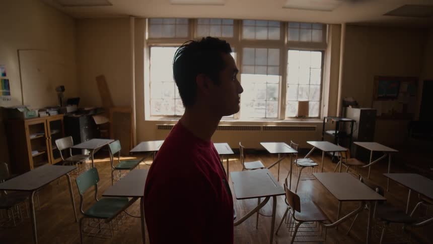 A man is standing in an empty school classroom staring ahead