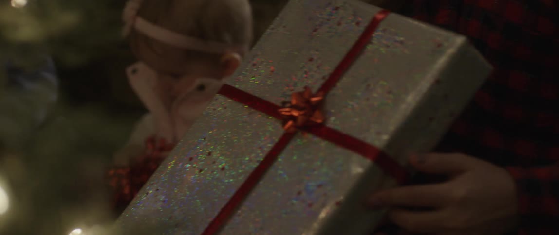 A young girl and toddler look at christmas gifts under the tree while kneeling in front of a fireplace