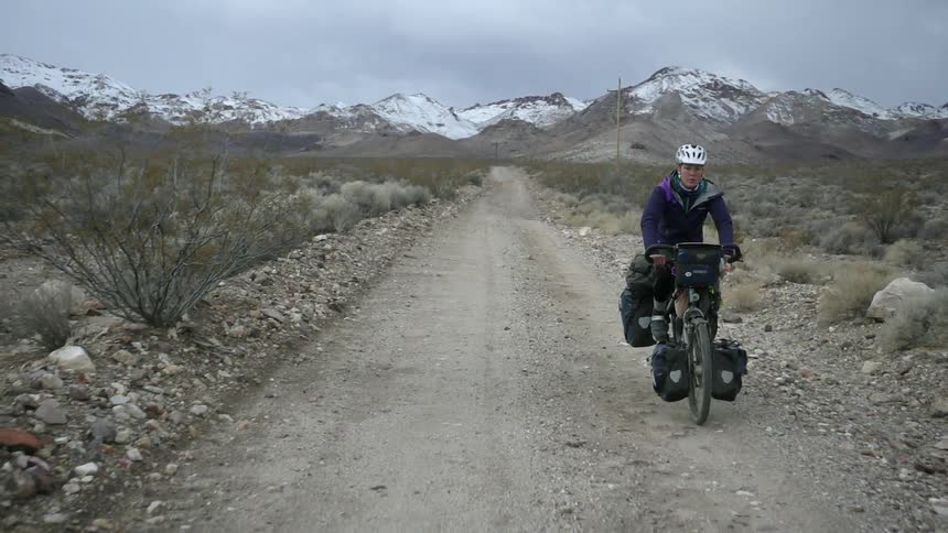 A man is riding his bike along a dirt road with snow covered mountains in the background