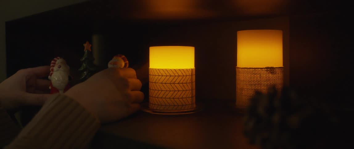 A white woman sets two christmas decorations on a shelf by lighted candles
