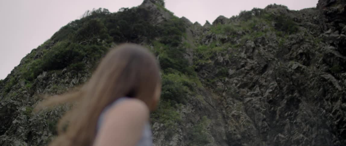 A woman stands near a rocky mountainside on a windy day