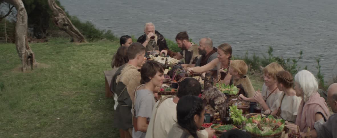 A large group of people dressed in costumes are having a meal next to a body of water