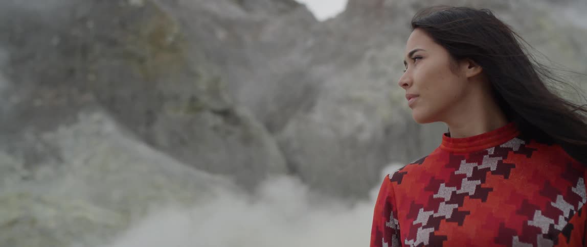 A woman stands in rocky mountains as smoke floats by