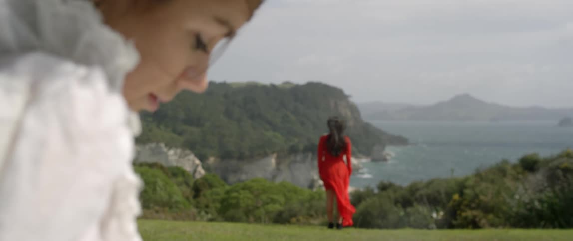 Two women, one in white and the other in red are standing in a grassy field overlooking the ocean