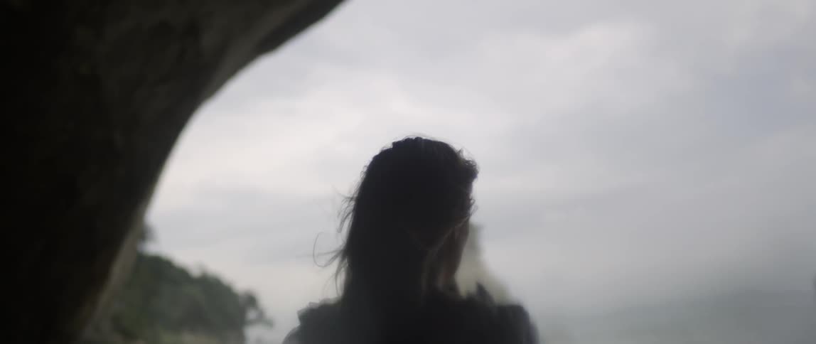 A woman stands along the shore as the wind blows her hair