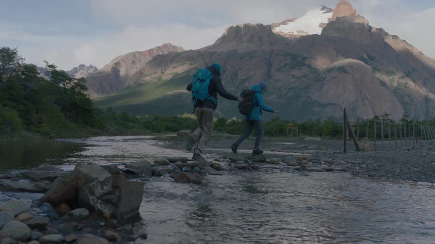 Two people are walking across a shallow stream with huge mountains in the background