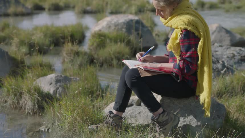 A woman sits on a rock and writes in a notebook