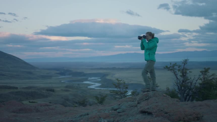 A woman in a teal jacket takes pictures near a winding river