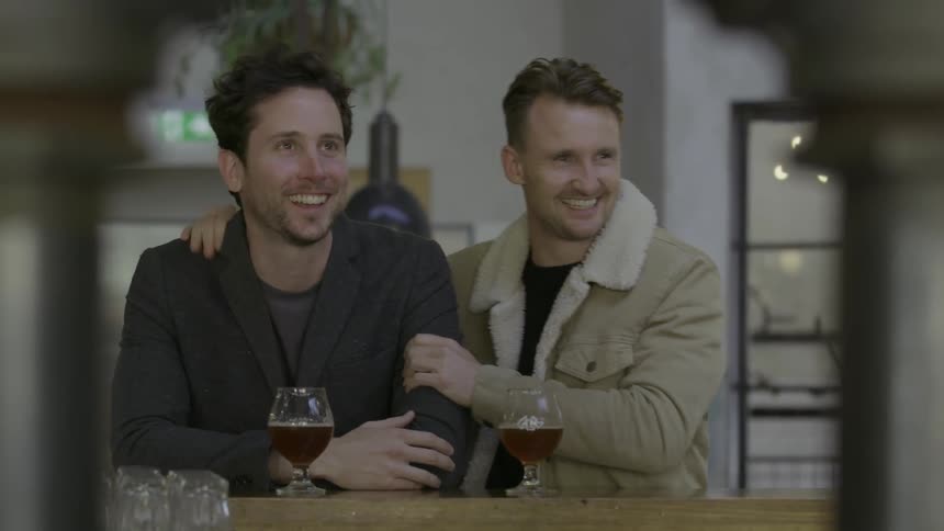 Two men sitting at a bar with beers talking and smiling