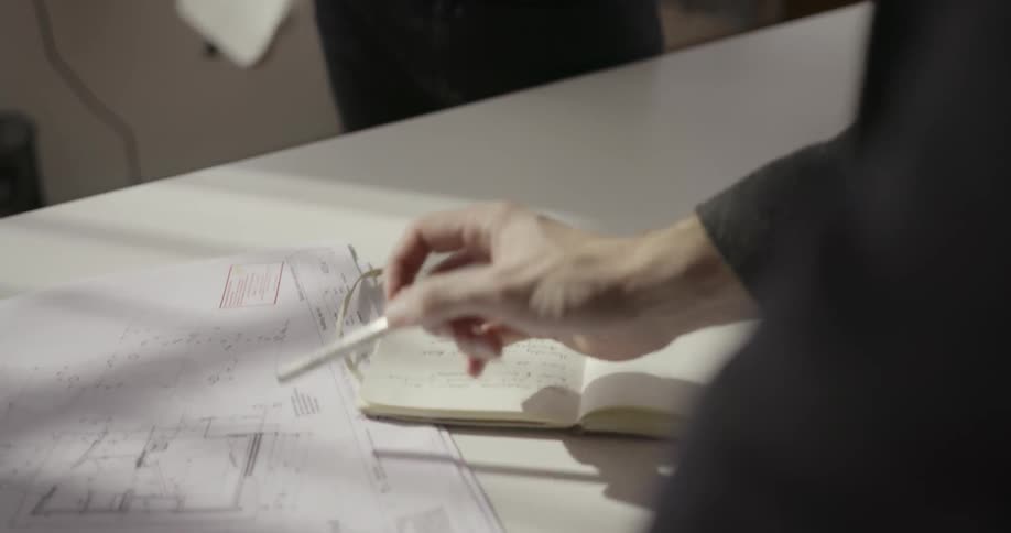 A man is writing in a book and another points towards a blueprint