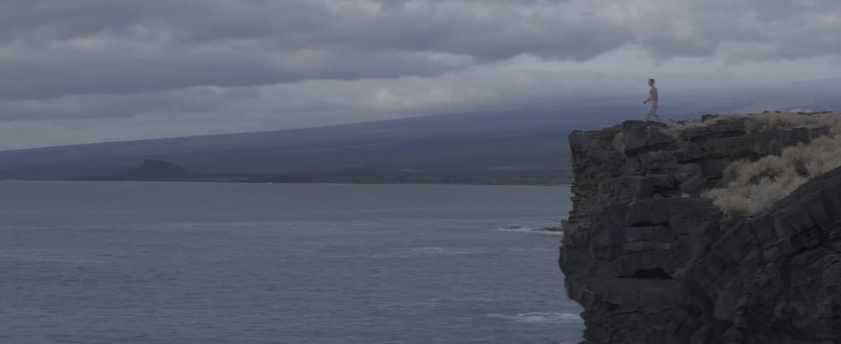 A man walks towards a cliff edge overlooking the water