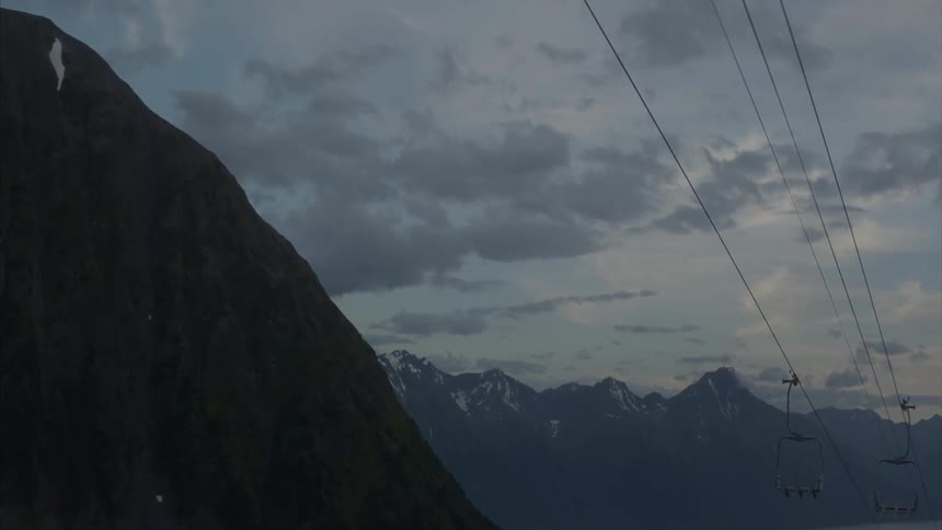 Chair lifts hanging from cables near mountains with snow on cloudy evening