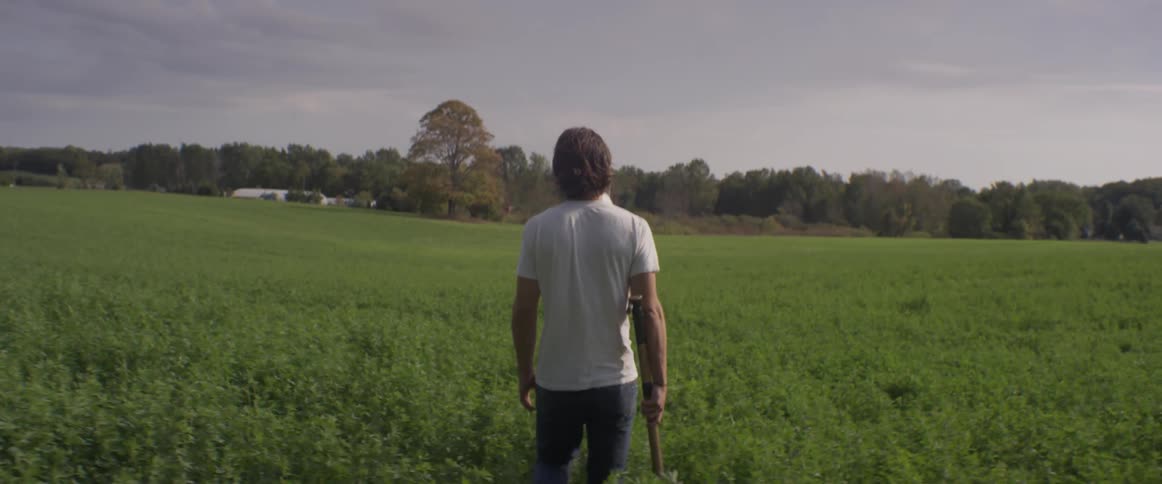 A man walks across a rural field with a hatchet