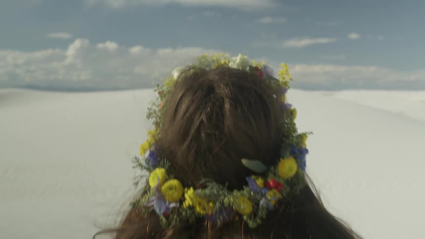 A girl wearing a flower crown is filmed from behind looking out over a cast desert