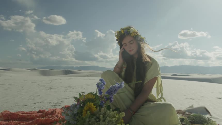 A woman wearing a flower crown sits on a cushion in the sand