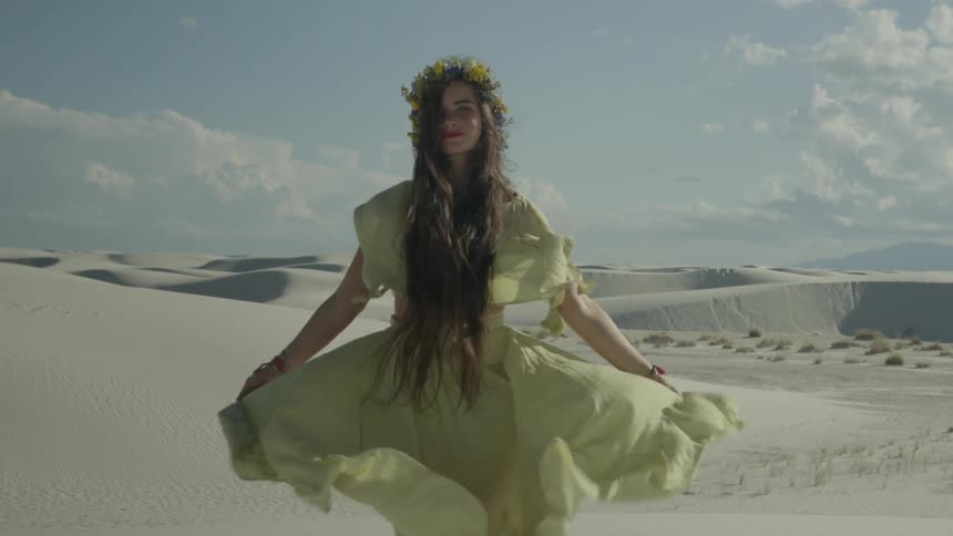 The wind blows a woman's dress as she stand in a sandy locale
