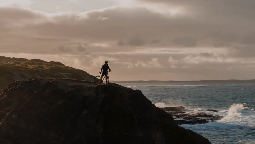 A person with a bike is standing on a cliff overlooking the ocean