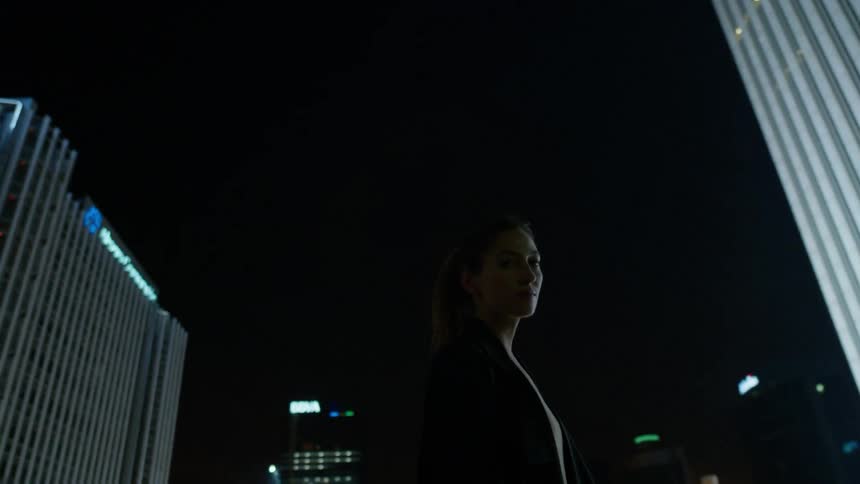 A woman stands in a city at night with lit buildings behind her