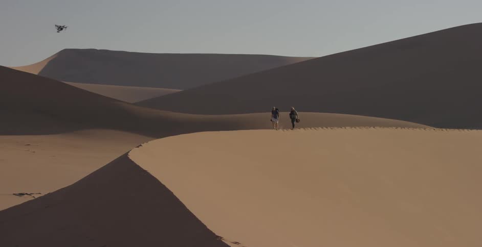 A drone is flying past two people walking on a sand dune
