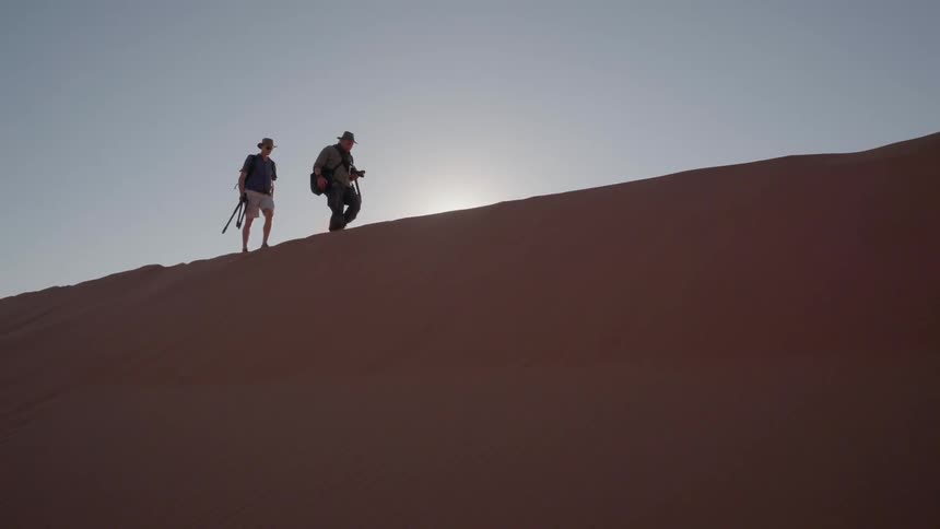 Men with camera walking on top of sand hill in dunes on sunny day