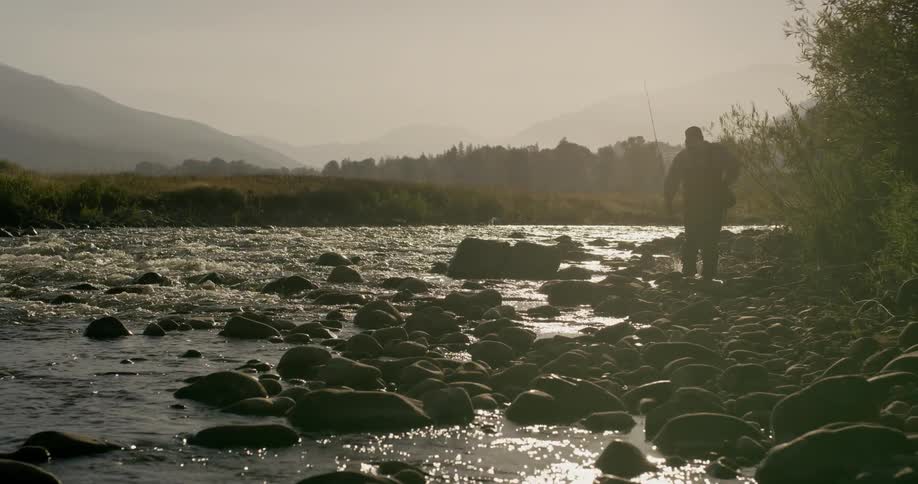 Man holding fishing pole is walking on rocky river shore