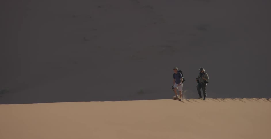 Two men carrying camera equipment are walking on a sand dune