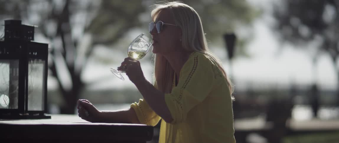 A woman sitting outside clinks her glass with another and takes a sip