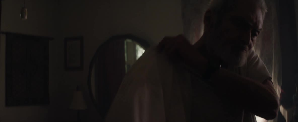 An elderly man puts on a button up shirt