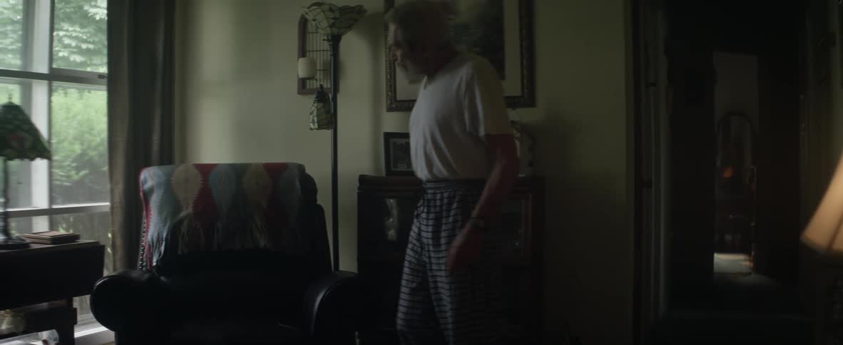 An elderly man walks towards and sits in a chair