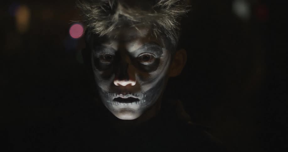 A young child in face paint stares with a light below his face