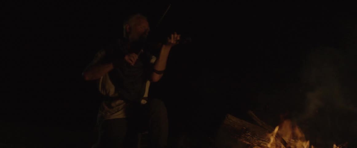 A man plays violin next to a campfire