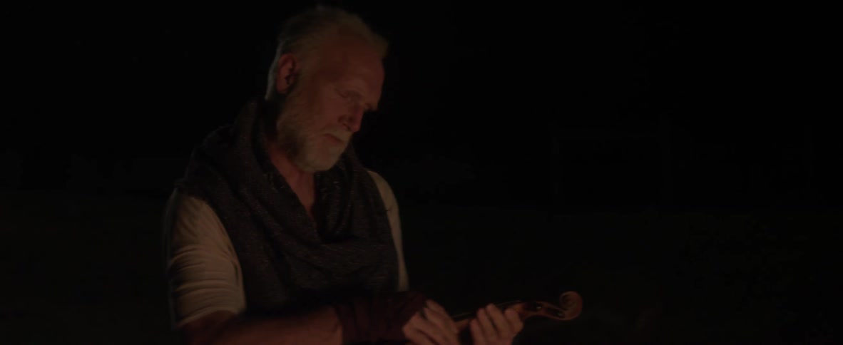 An elderly man holds a violin while a campfire burns