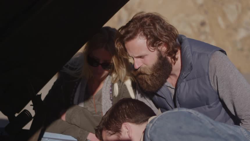 A heavily bearded man, along with a woman and another male, check under the hood of a car