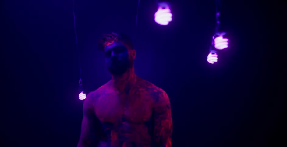 Man in body paint stands under several spiral blacklight bulbs