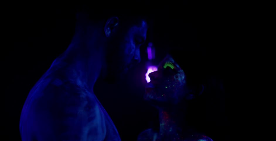 A man and woman in glowing body paint stare at each other